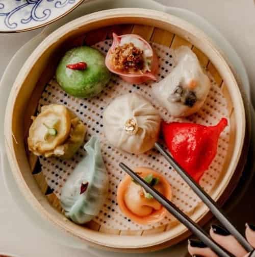Green, red, white, yellow and orange dumplings are in the steamer, and a pair of chopsticks are sandwiching the dumplings.