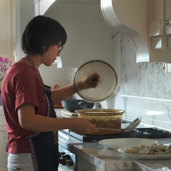 A woman in a red T-shirt and a blue apron is standing by the stove making 