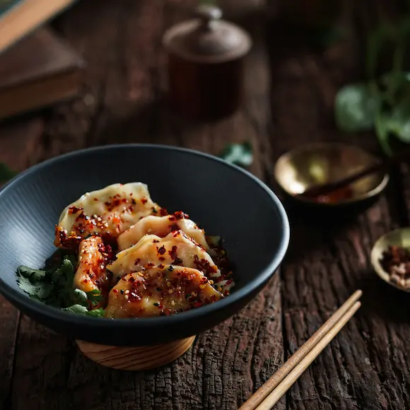On the wooden table sits a black bowl, which contains dumplings with chili